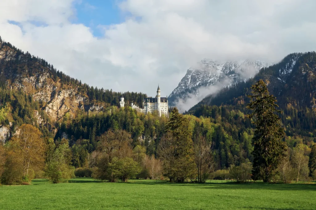 In the image, the majestic Neuschwanstein Castle stands nestled on a forested hillside surrounded by lush green valleys and dense trees. Majestic, snow-capped mountains rise in the background under a partly cloudy sky, framing the fairytale-like landscape.