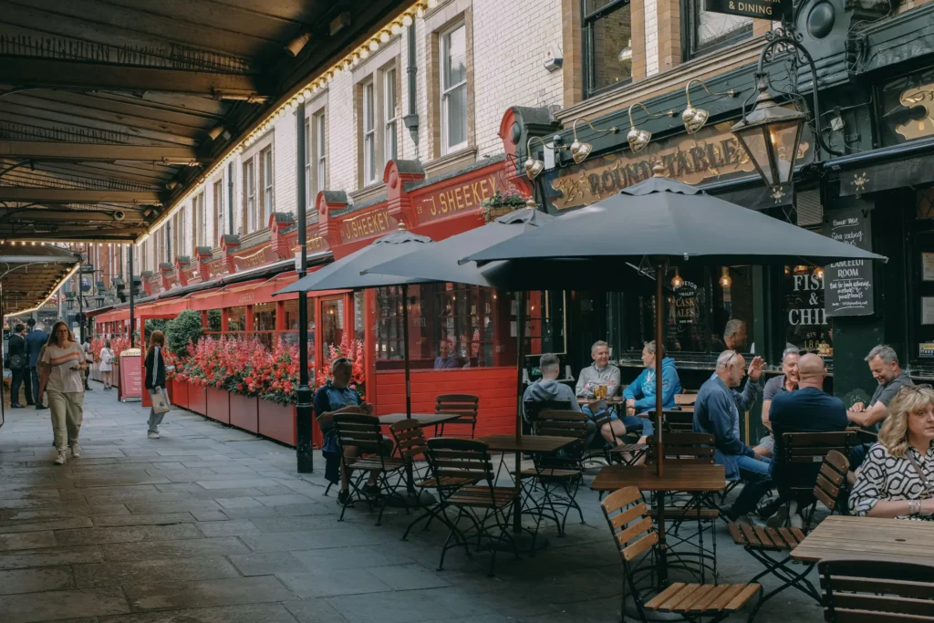 The image shows a lively street scene featuring outdoor patio seating under large dark umbrellas outside "The Round Table" pub and "J. Sheekey" restaurant. Patrons are seated at wooden tables enjoying drinks, while pedestrians walk along the covered stone sidewalk next to vibrant red flowerbeds.