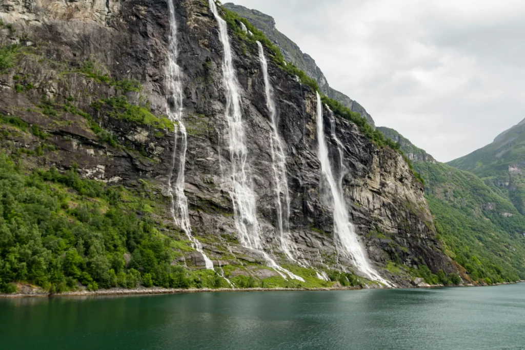 A series of long, thin waterfalls known as the Seven Sisters cascade down a steep, rocky cliffside covered with patches of green vegetation. The water flows directly into the calm, emerald-green waters of a fjord under an overcast sky.