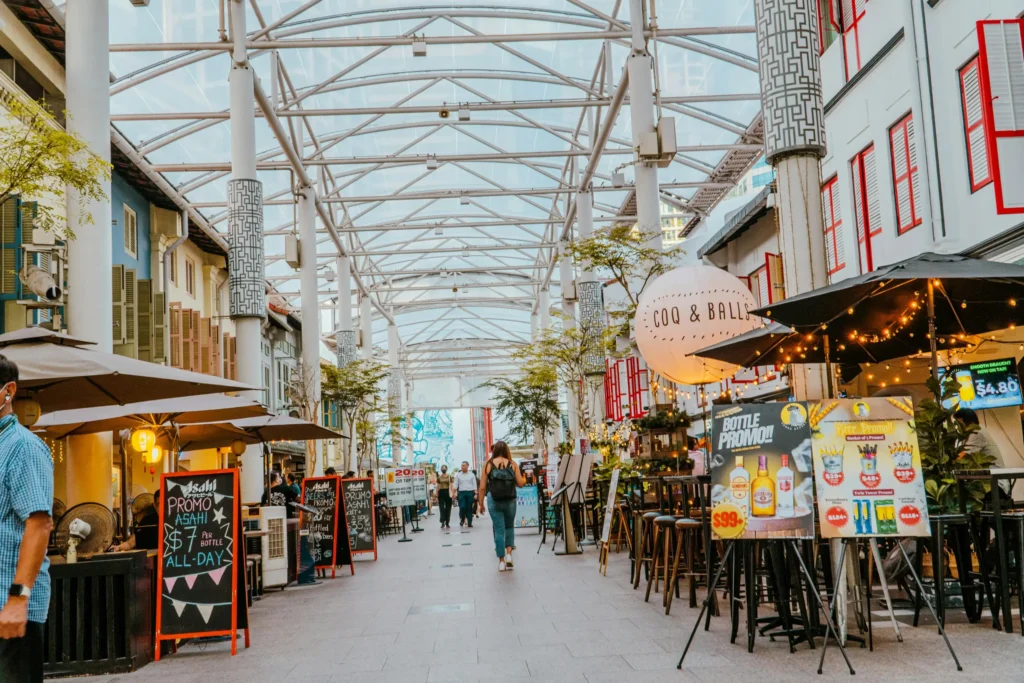 A lively pedestrian walkway is sheltered by a modern glass and steel canopy and lined with colorful shuttered buildings, outdoor bar seating, and promotional signboards. People stroll down the center of the path, which features a whimsical round balloon sign reading "COQ & BALLS" illuminated by string lights.