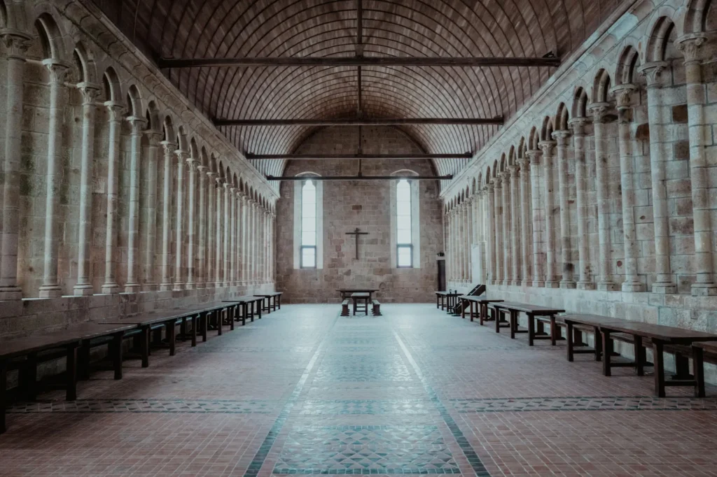 An expansive, historic stone hall or refectory features a high arched wooden ceiling, tiled floors, and symmetrical rows of columns lining both sides. Long wooden tables and benches stretch along the walls, leading toward a stone wall at the far end adorned with a simple wooden cross between two tall, narrow windows.