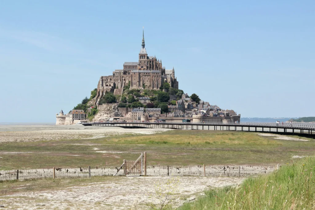 A panoramic, wide-angle shot shows the historic, tidal island of Mont Saint-Michel rising majestically under a clear blue sky in Normandy, France. In the foreground, a modern pedestrian bridge stretches across a vast, grassy mudflat landscape, leading directly to the fortified medieval village and abbey.