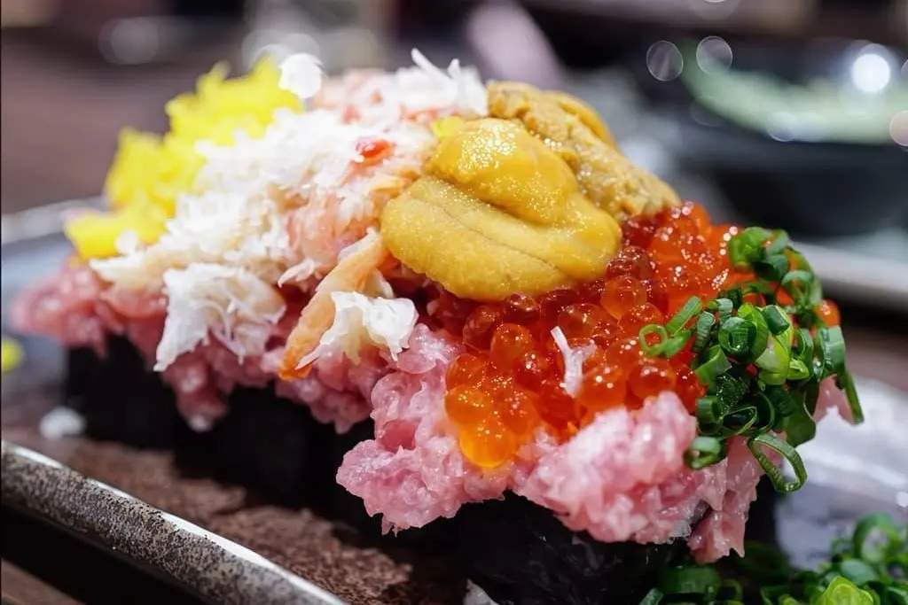 A close-up of the dish shows sushi rolls heavily piled with a variety of colorful seafood toppings. The vibrant mound includes minced pink tuna, glistening orange salmon roe, bright yellow sea urchin (uni), shredded crabmeat, chopped green onions, and yellow pickled radish.