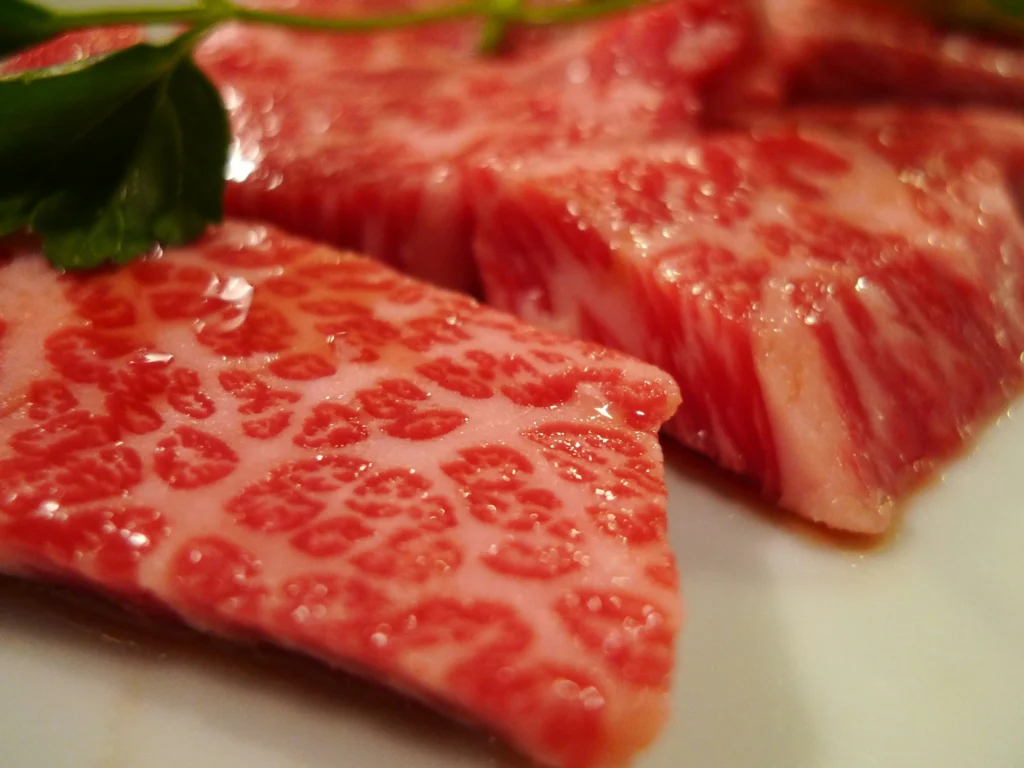 A close-up shot displays raw slices of highly marbled Wagyu beef, showcasing an intricate web of white fat distributed throughout the pink meat. A single green leaf rests on top of the meat against a soft-focus white background.