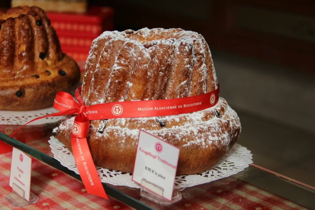 A traditional, ring-shaped Kougelhopf cake sits on a decorative paper doily on a glass shelf, dusted with powdered sugar and wrapped in a bright red ribbon labeled "Maison Alsacienne de Biscuiterie." A small price tag reading "Kougelhopf Traditionnel" stands in front of the pastry, while another bundt-style cake is partially visible in the blurred background.