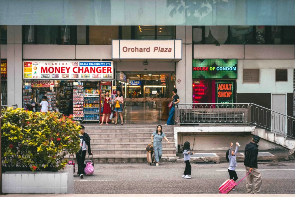 Candid street scene outside the entrance of Orchard Plaza in Singapore, featuring pedestrians, children playing with pink luggage, and signs for a money changer and adult shop.