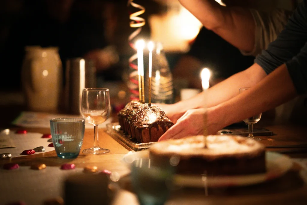 Hands gently place a chocolate loaf cake with three lit birthday candles onto a dimly lit table surrounded by glasses and party decorations. The warm, glowing scene captures a cozy, intimate celebration with friends and family.