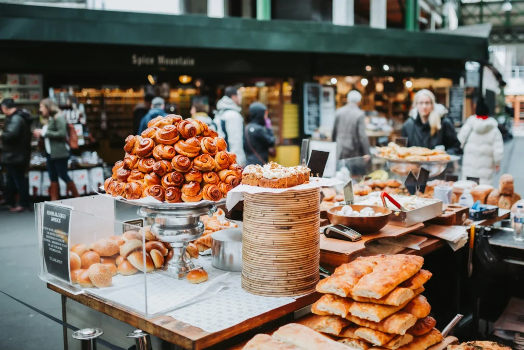 The image displays a vibrant outdoor market stall overflowing with fresh pastries and artisanal breads. A massive, towering pyramid of golden-brown cinnamon rolls serves as the focal point, while blurred shoppers stroll past storefronts in the background.
