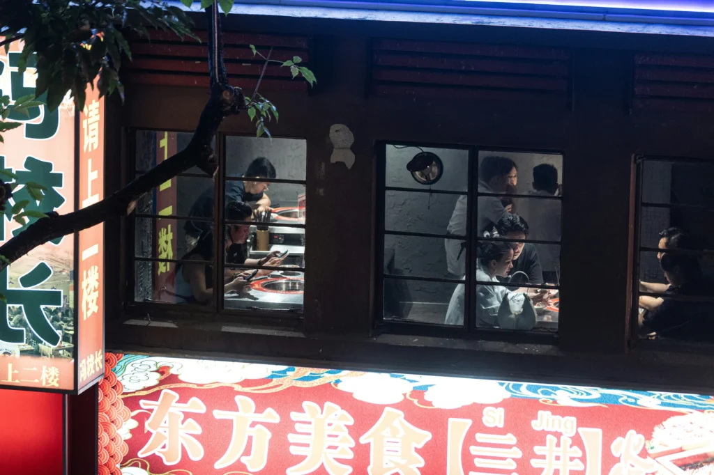 An elevated, nighttime view looks through the windows of a brightly lit restaurant where diners are seated at tables. Below the windows, a prominent, colorful Chinese sign glows with stylized text and patterns.