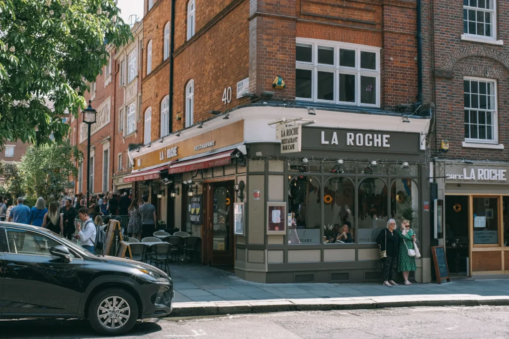 The image captures a bustling street corner featuring a multi-story brick building that houses the "La Roche" restaurant and bar. Pedestrians walk along the sidewalk and patrons sit at outdoor tables, while a black SUV is parked in the foreground on a sunny day.