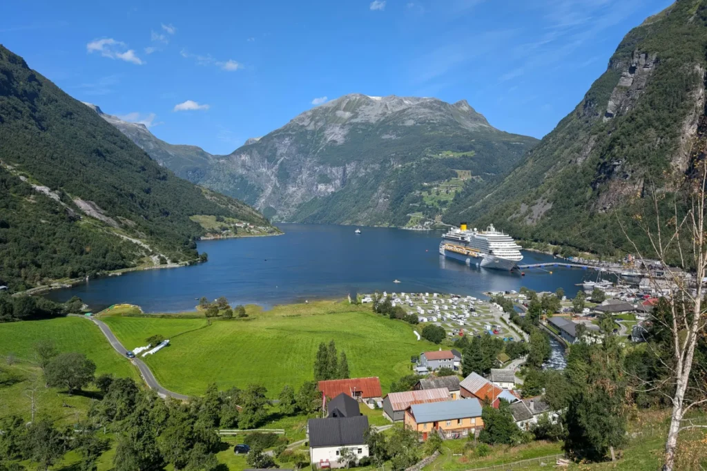 The image captures a scenic view of a deep blue fjord nestled between towering, lush green mountains under a clear sky. A large white cruise ship is docked near a small coastal village with colorful houses, green fields, and a camper park lining the shore.