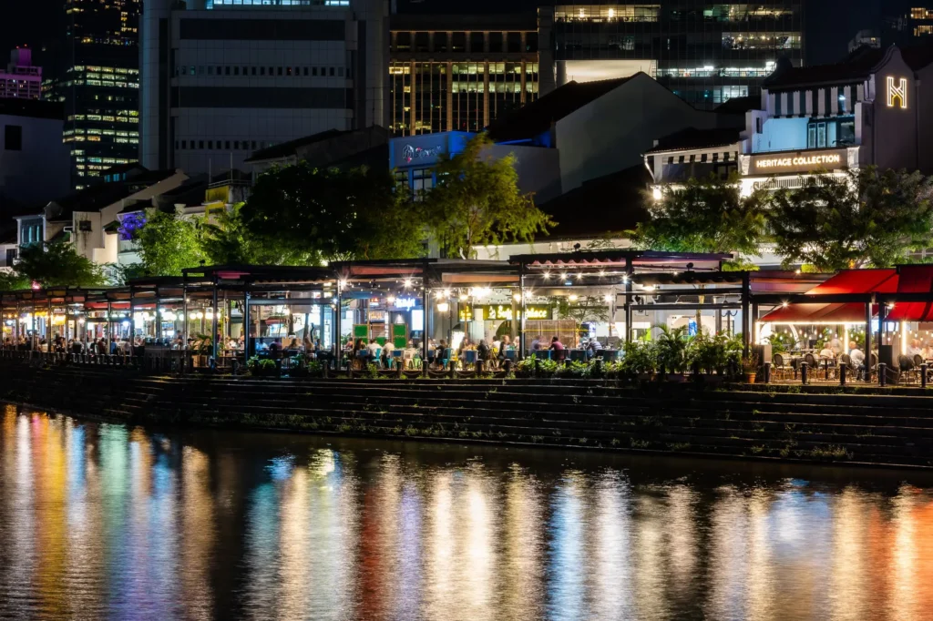 A vibrant, brightly lit row of outdoor restaurants and bars lines a waterfront at night, with patrons seated under canopies and trees. The colorful lights from the venues cast shimmering reflections across the dark surface of the river, while modern high-rise buildings loom in the background.