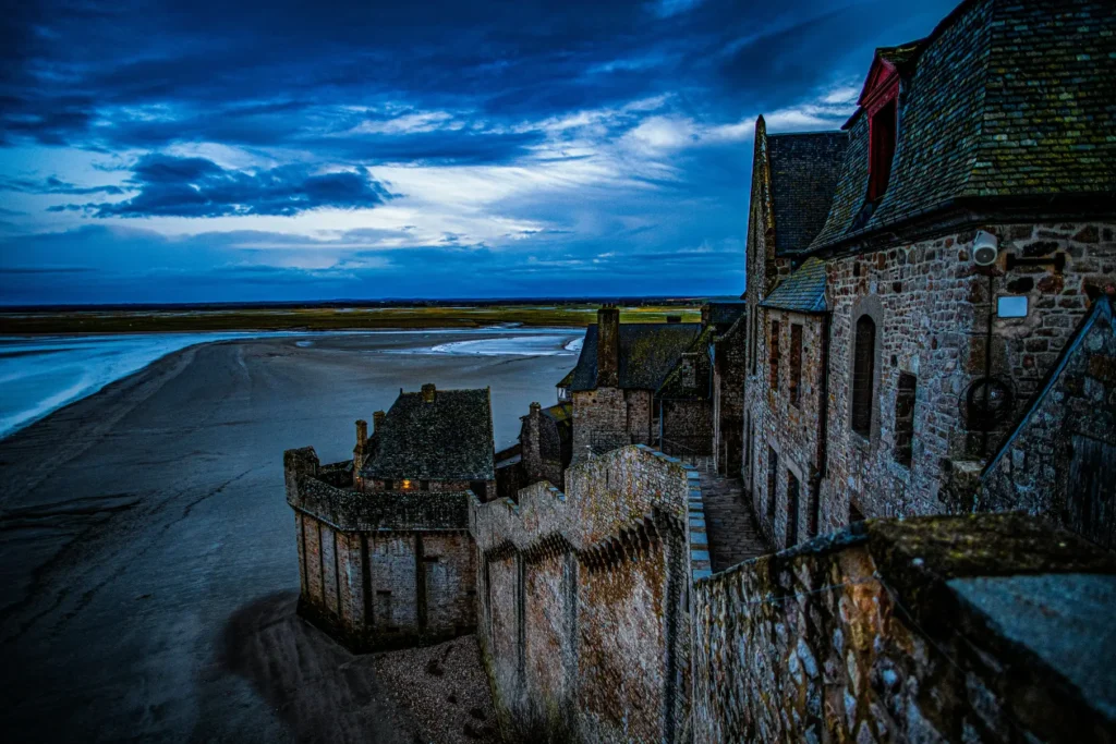 A high-angle shot captures the stone walls and buildings of Mont Saint-Michel overlooking a vast mudflat under a dramatic, moody blue sky. The ancient stone structures stretch along the right side of the frame, contrasting with the wide, empty landscape below during low tide.