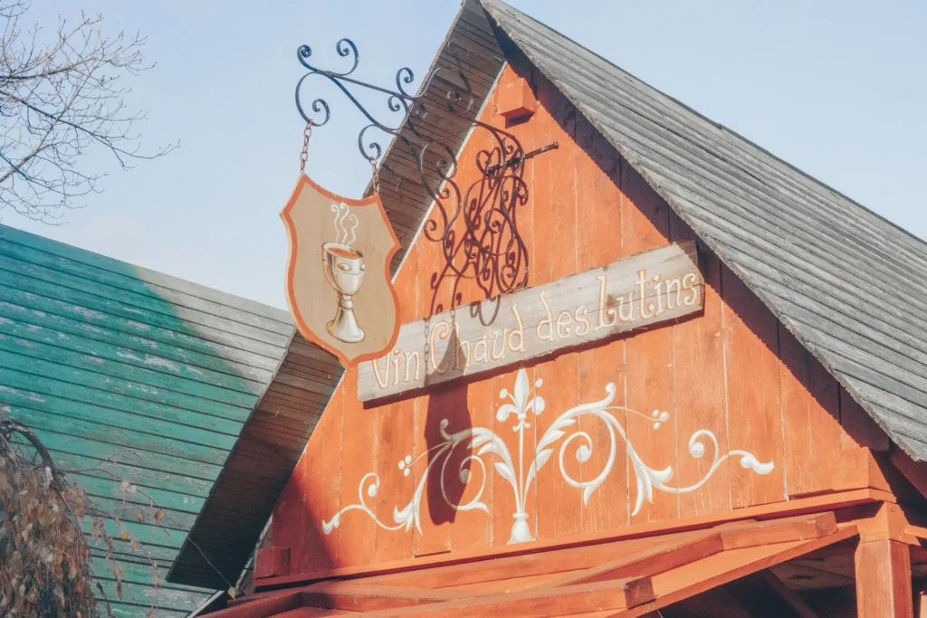 A rustic wooden sign reading "Vin Chaud des lutins" is mounted on the orange-painted facade of a building with a steep, weathered roof. To the left, a shield-shaped hanging sign displays an illustration of a steaming goblet, all set against a clear blue sky.