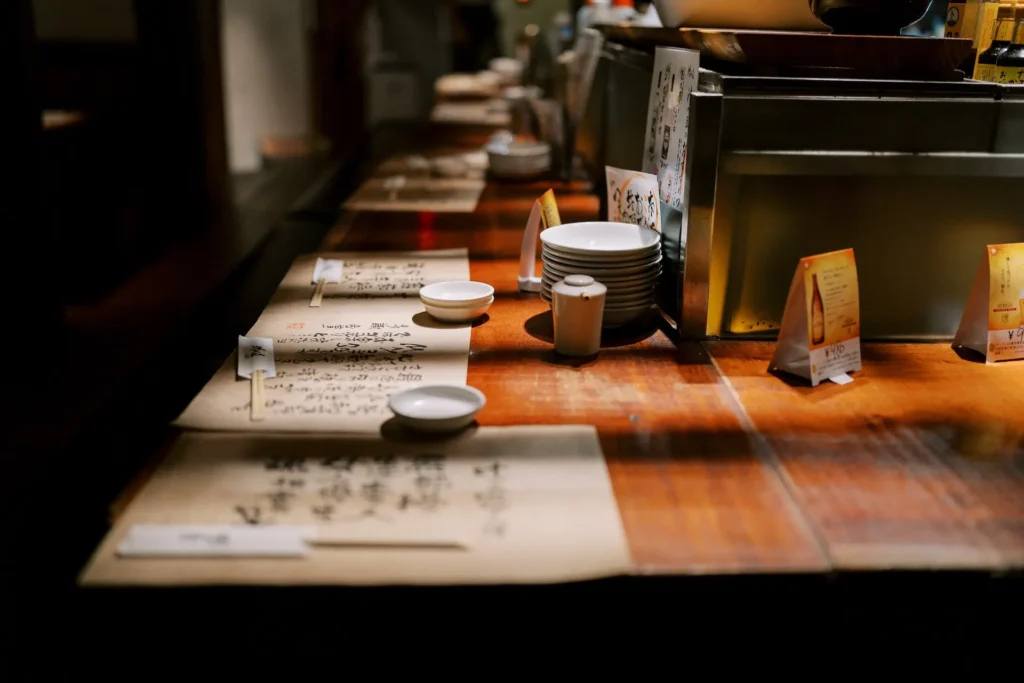 The image shows a dimly lit restaurant counter set with small plates, chopsticks, and menus featuring Japanese calligraphy. Warm light illuminates the long wooden counter, creating a cozy and inviting dining atmosphere.