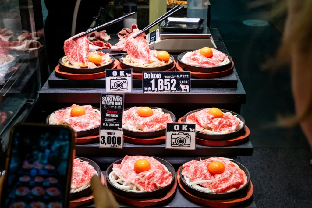 A display features multiple plates of raw, heavily marbled Wagyu beef served over noodles and topped with a raw egg yolk. Small signs indicate the pricing for "Deluxe" and "Super Deluxe" options, while a person captures a photo of the food on their smartphone in the foreground.