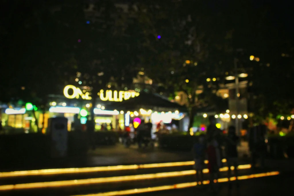 A blurry, out-of-focus night shot shows a brightly lit outdoor venue with a large glowing sign that appears to read "ONE FULLERTON." In the foreground, the faint silhouettes of people stand near steps illuminated by parallel lines of warm light.