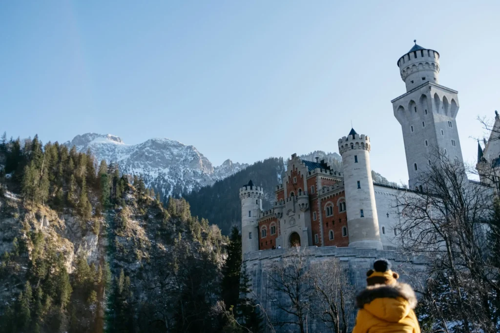 A large, historic castle with high stone towers and red brick accents stands prominently against a backdrop of steep, pine-covered mountain slopes and snow-capped peaks. In the foreground, a person wearing a yellow winter coat with a fur-trimmed hood is seen from behind, looking up at the majestic fortress under a clear blue sky.