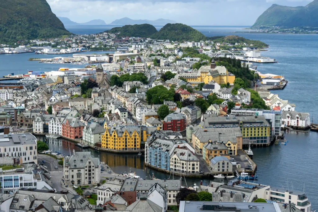 An aerial view of a coastal city shows tightly packed, colorful buildings with distinct grey roofs lining a system of narrow waterways and harbors. Lush green hills and distant mountains frame the dense urban landscape under a partly cloudy sky.