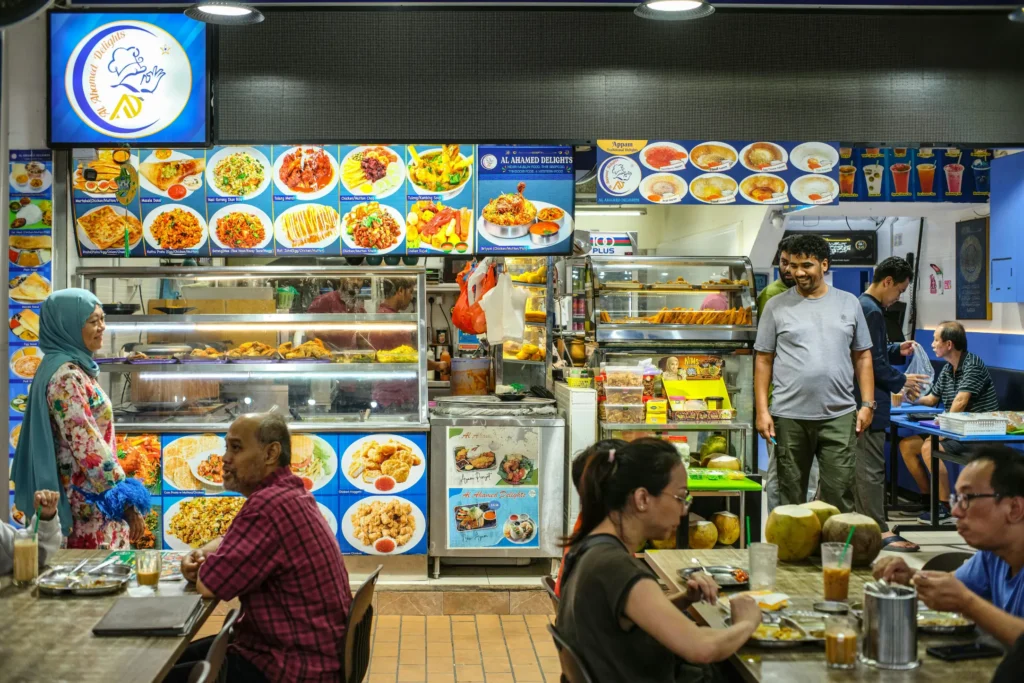 A bustling hawker stall or casual eatery named "Al Ahamed Delights" displays a vibrant menu with pictures of various dishes above the food counter. In the foreground, customers are seated at wooden tables enjoying their drinks and meals while stall workers and other patrons stand nearby.