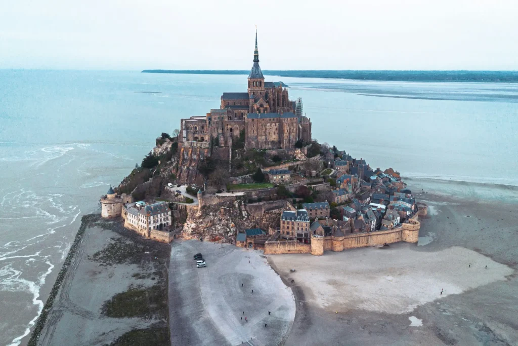 An aerial, high-angle view captures the historic tidal island of Mont-Saint-Michel in Normandy, France, surrounded by vast, shimmering mudflats and a calm sea under an overcast sky. The dramatic gothic abbey sits at the peak of the rocky island, with stone ramparts and medieval village buildings clustering tightly around its base.
