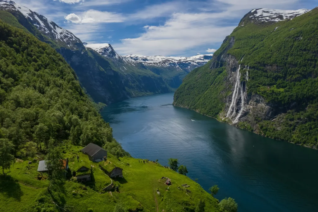 An aerial view captures a deep blue fjord winding between steep, lush green mountains topped with patches of snow. In the foreground, a grassy hillside features a few small, sod-roofed wooden cabins, while a dramatic multi-stream waterfall cascades down the rugged cliffs on the right.