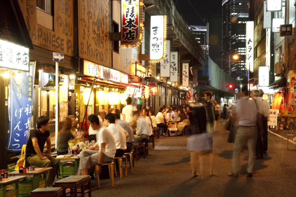 A bustling Tokyo alleyway at night features patrons dining outside under glowing izakaya signs and lanterns. Pedestrians walk down the narrow street, capturing a lively and vibrant nightlife scene in Japan.