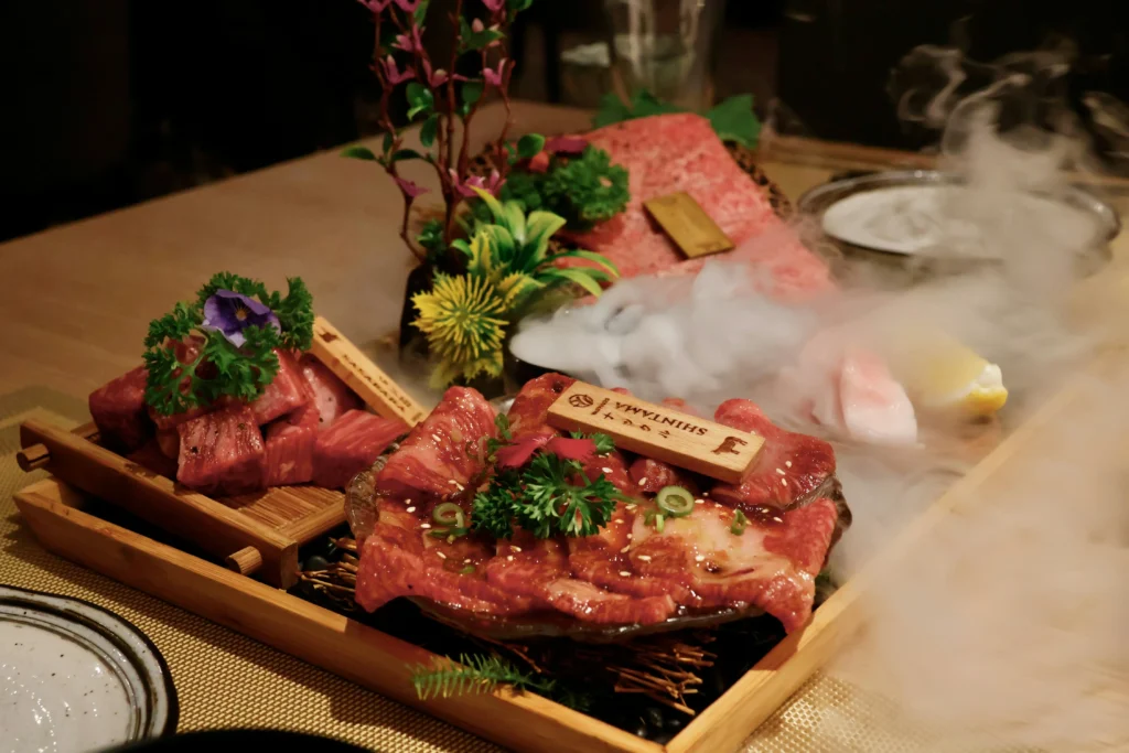 A premium assortment of raw wagyu beef is beautifully arranged on a wooden platter, decorated with fresh herbs, flowers, and a dramatic dry ice fog effect. The thinly sliced and cubed meats feature rich marbling and are accompanied by small wooden labels indicating the cuts.