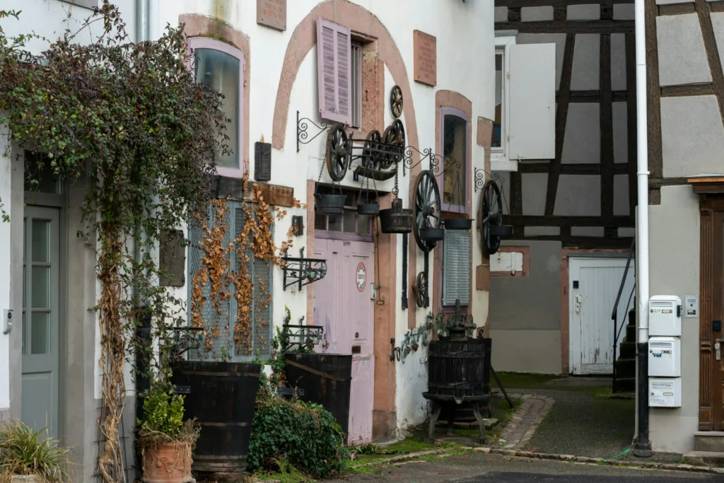 A charming, narrow alleyway features old-fashioned European buildings decorated with rustic wooden wheels, metal chains, and hanging buckets. Overgrown green and dried vines scale the white walls next to a light pink door, while a timber-framed building stands in the background.
