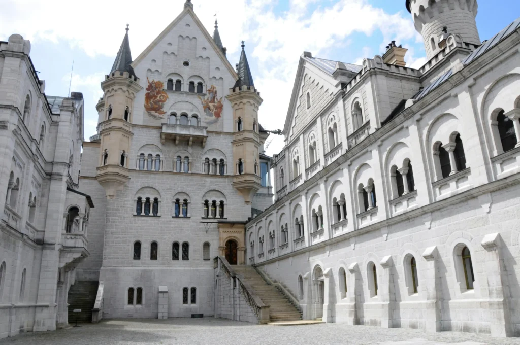 A bright, eye-level view captures the stone courtyard of Neuschwanstein Castle, surrounded by white limestone walls with arched windows and pointed turrets. An outdoor staircase leads to an arched doorway on the main building, which features two colorful frescoes painted high on its facade.