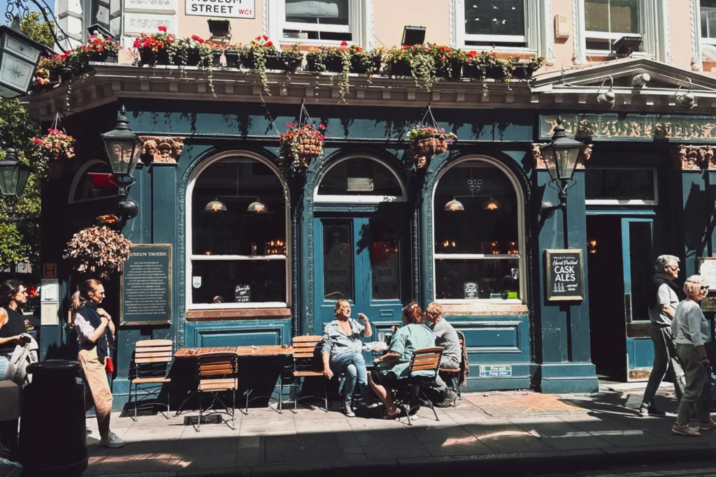 An exterior view of the historic Museum Tavern pub features its dark green facade, large arched windows, and vibrant flower boxes lining the upper ledge. In front of the establishment, patrons sit at wooden tables on the sunlit sidewalk while pedestrians walk by on either side.
