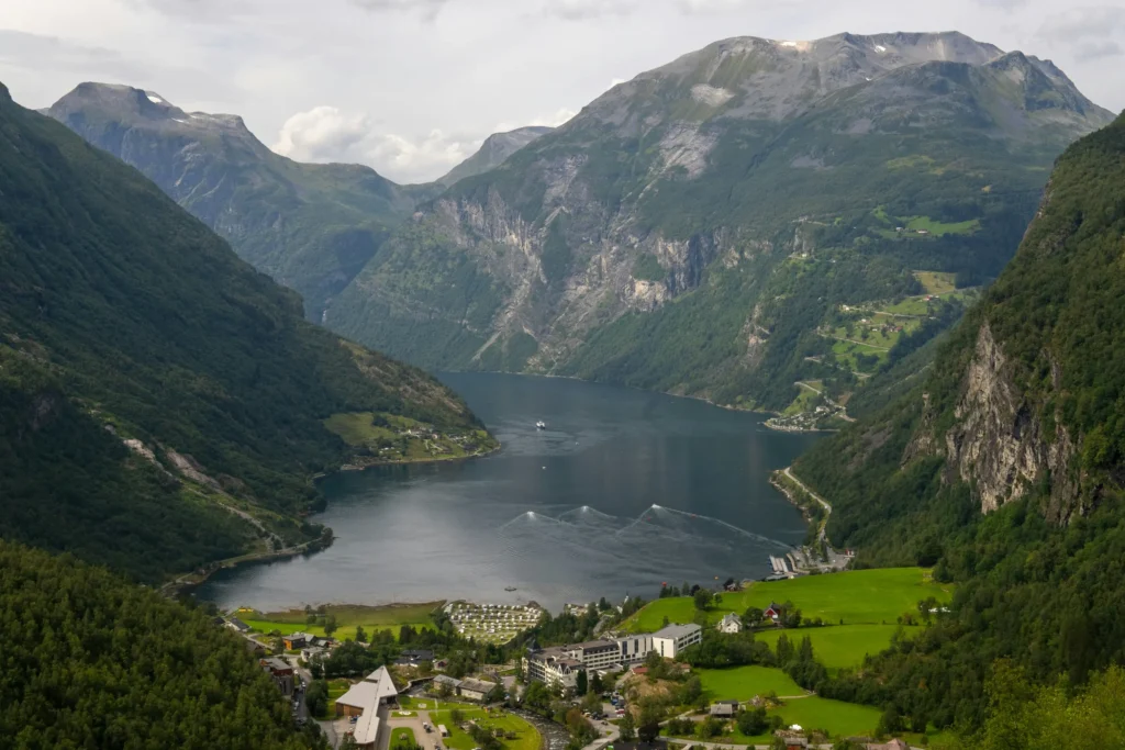 An elevated, wide-angle view captures a deep blue fjord nestled between steep, lush green mountains under a slightly overcast sky. At the base of the valley, a small village with green fields, buildings, and a campground sits along the waterfront as boats leave wake trails across the water.