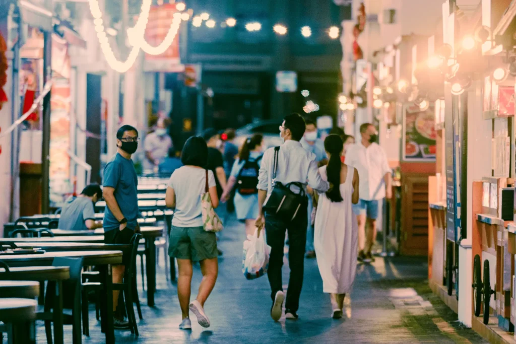 A crowded outdoor street at night is illuminated by warm overhead string lights and bright storefront displays, with people walking along the path and dining at tables. In the foreground, several pedestrians are seen from behind, including a couple walking side-by-side, while a man in a blue shirt and black face mask looks toward the camera.