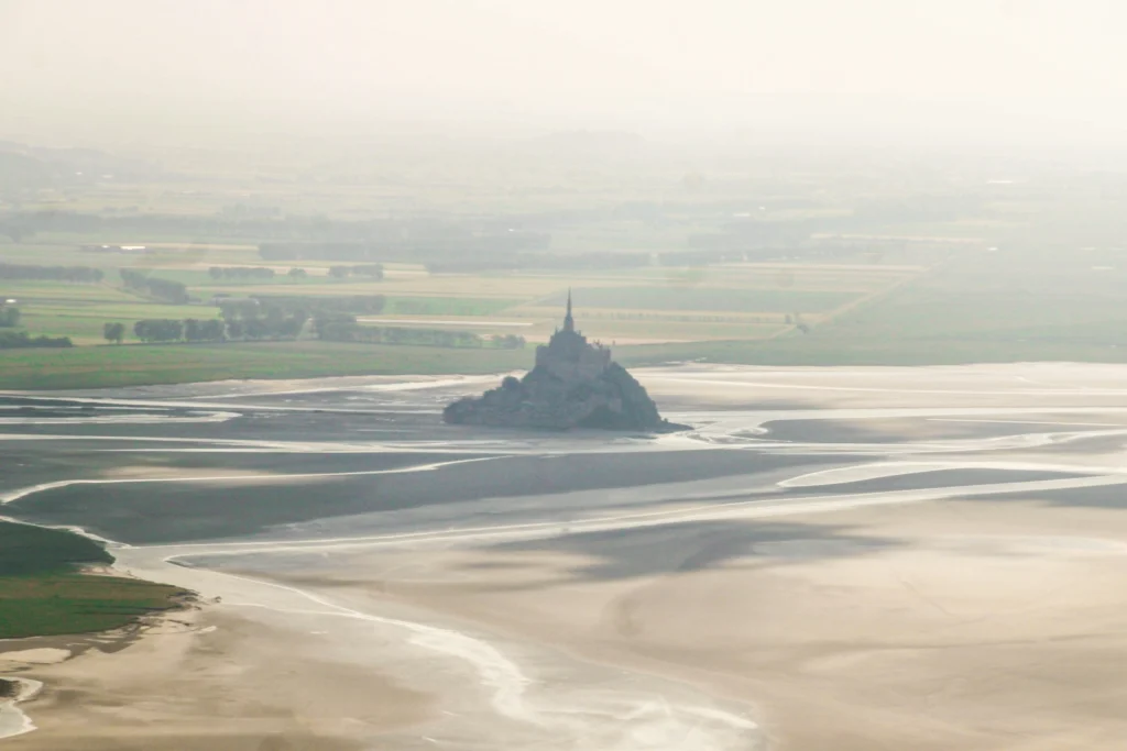 An aerial, hazy view captures the silhouette of Mont-Saint-Michel rising from the surrounding tidal flats and shallow waters. In the background, a flat landscape of green and yellow agricultural fields stretches into the misty distance.