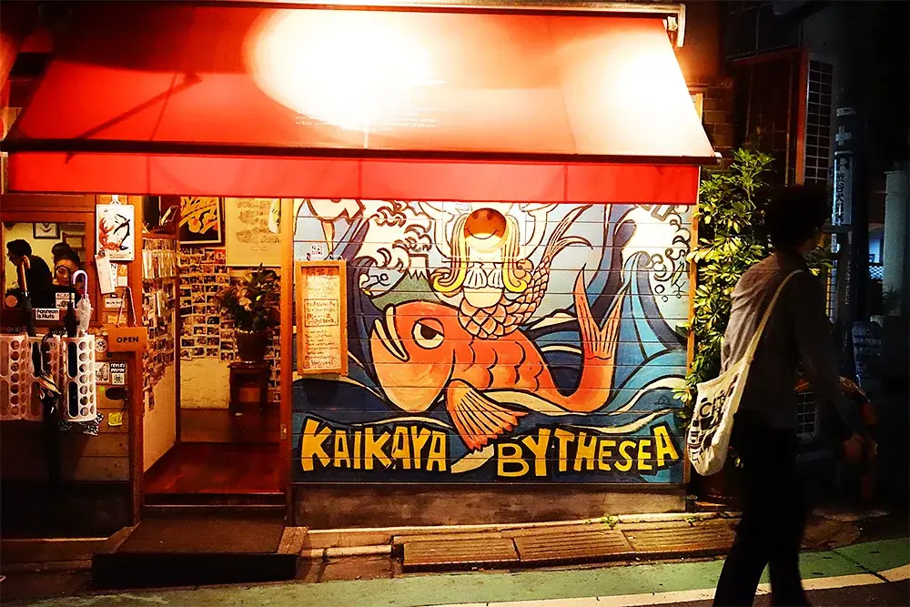 A vibrant storefront for a restaurant named "Kaikaya by the Sea" features a large, colorful mural of a mermaid riding a large orange fish amidst blue ocean waves. A glowing red awning illuminates the entrance at night while a pedestrian with a shoulder bag walks past on the street in front of image-asset.webp.