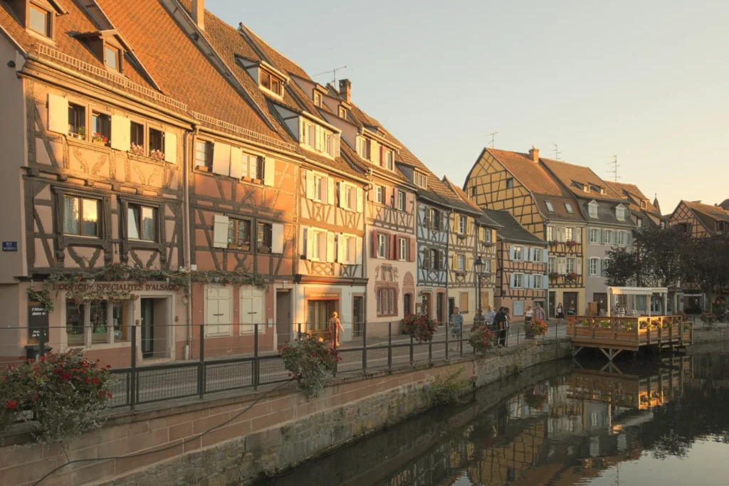 The image features a row of historic, half-timbered buildings with steep roofs lined along a calm canal during the warm glow of late afternoon. People stroll along the fenced walkway beside the water, which beautifully reflects the architecture and a small wooden outdoor dining deck.