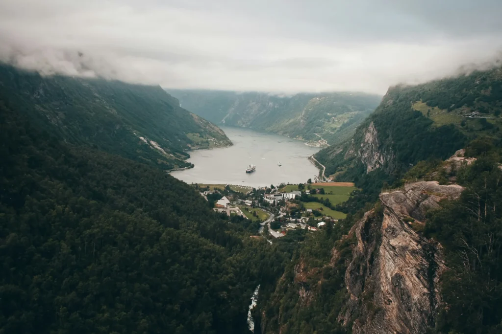 An elevated, wide-angle view captures a dramatic fjord nestled between steep, lush green mountains partially shrouded in low-hanging clouds. In the valley below, a small coastal village sits at the edge of the water where a cruise ship and a boat are anchored.