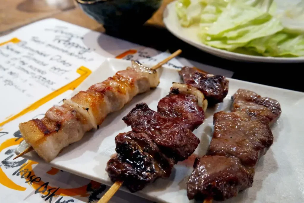 The image features three skewers of grilled meat served on a white square plate. A side plate of green cabbage leaves and a handwritten menu are visible in the background on the wooden table.