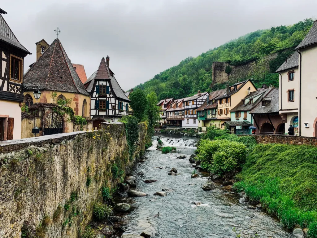 A scenic view shows a shallow, rocky river flowing through a charming European village lined with traditional half-timbered houses. A weathered stone wall borders the left bank, while a lush green hillside rises under an overcast sky in the background.