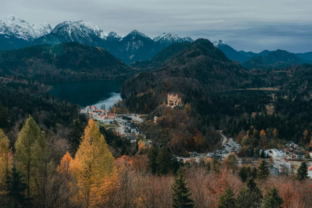 An elevated, wide shot of a valley shows Hohenschwangau Castle nestled among dense autumn forests next to a deep blue lake. In the background, majestic snow-capped alpine mountains rise against an overcast, moody sky.
