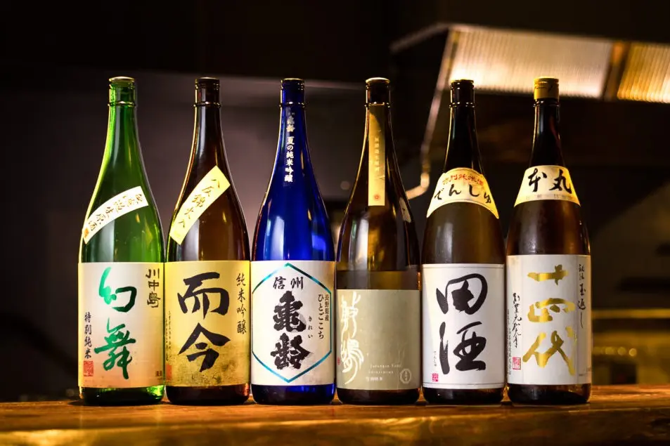 In the image, a row of six distinct Japanese sake bottles stands side-by-side on a dark wooden counter against a dimly lit background. The bottles feature a variety of glass colors—including green, brown, and vibrant blue—each adorned with traditional white labels displaying bold Japanese calligraphy.
