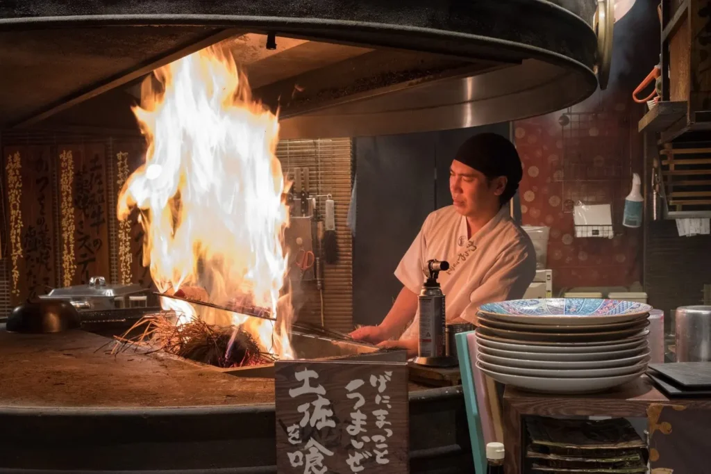 In the image, a chef in a white uniform and black head covering watches a large, vibrant burst of flames erupting from a straw-fired grill station. The setting is a traditional Japanese restaurant kitchen, complete with stacked ceramic plates, a blowtorch, and wooden signs with Japanese text.