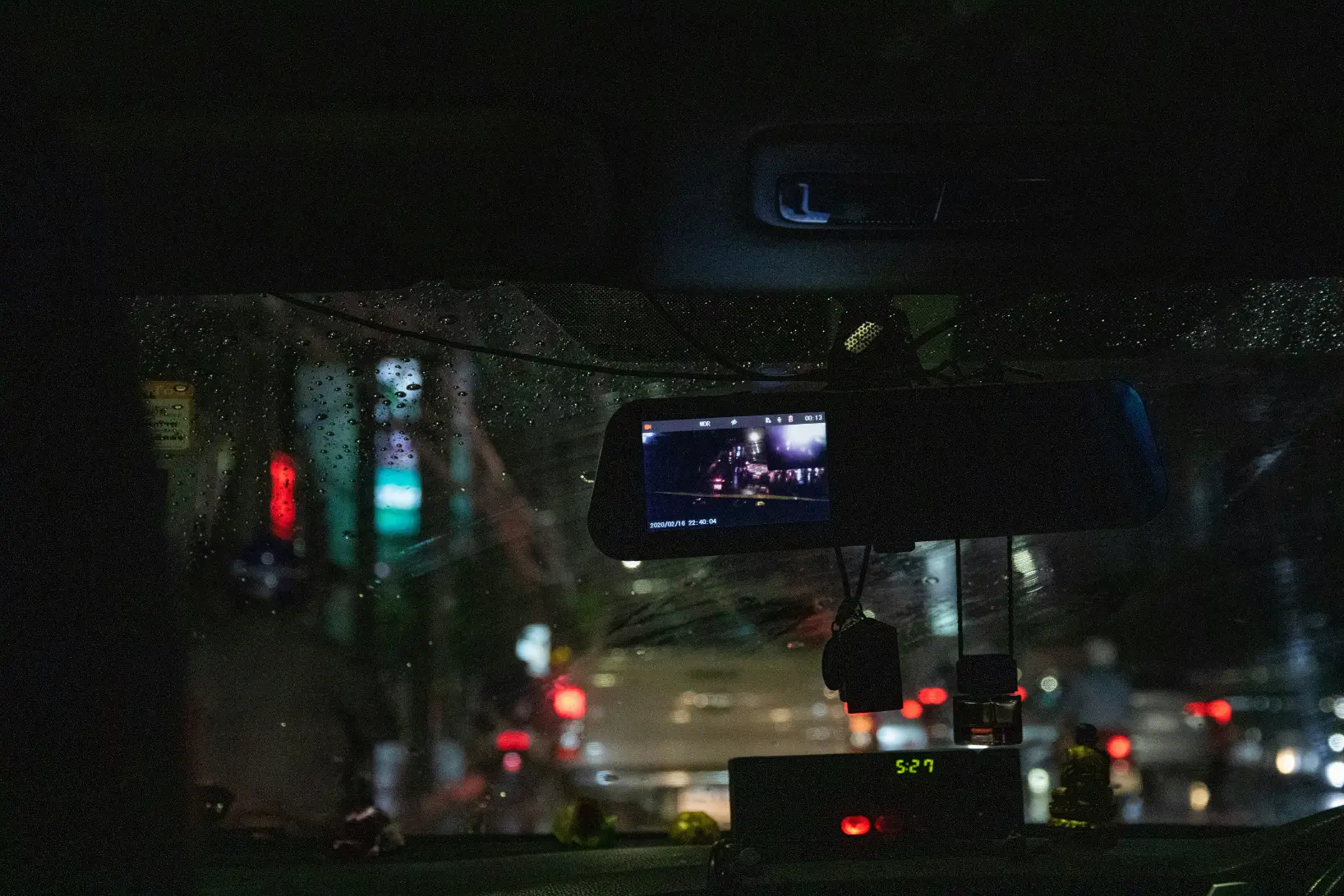 The image shows the interior of a dark vehicle looking out through a rain-covered windshield at a blurry city street at night. A rearview mirror with a built-in dashcam display is visible in the center, showing a small live feed of the road ahead amidst the ambient city lights.