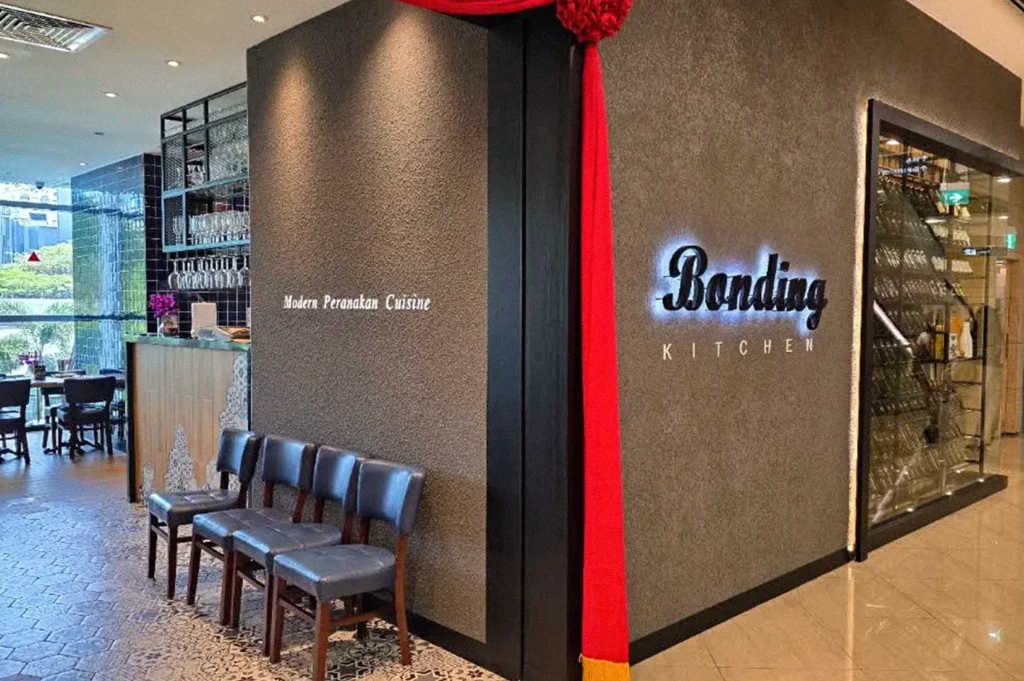 Entrance to a modern Peranakan restaurant with a dark textured wall, illuminated “Babatin Kitchen” sign, red decorative drapery, and a row of chairs.