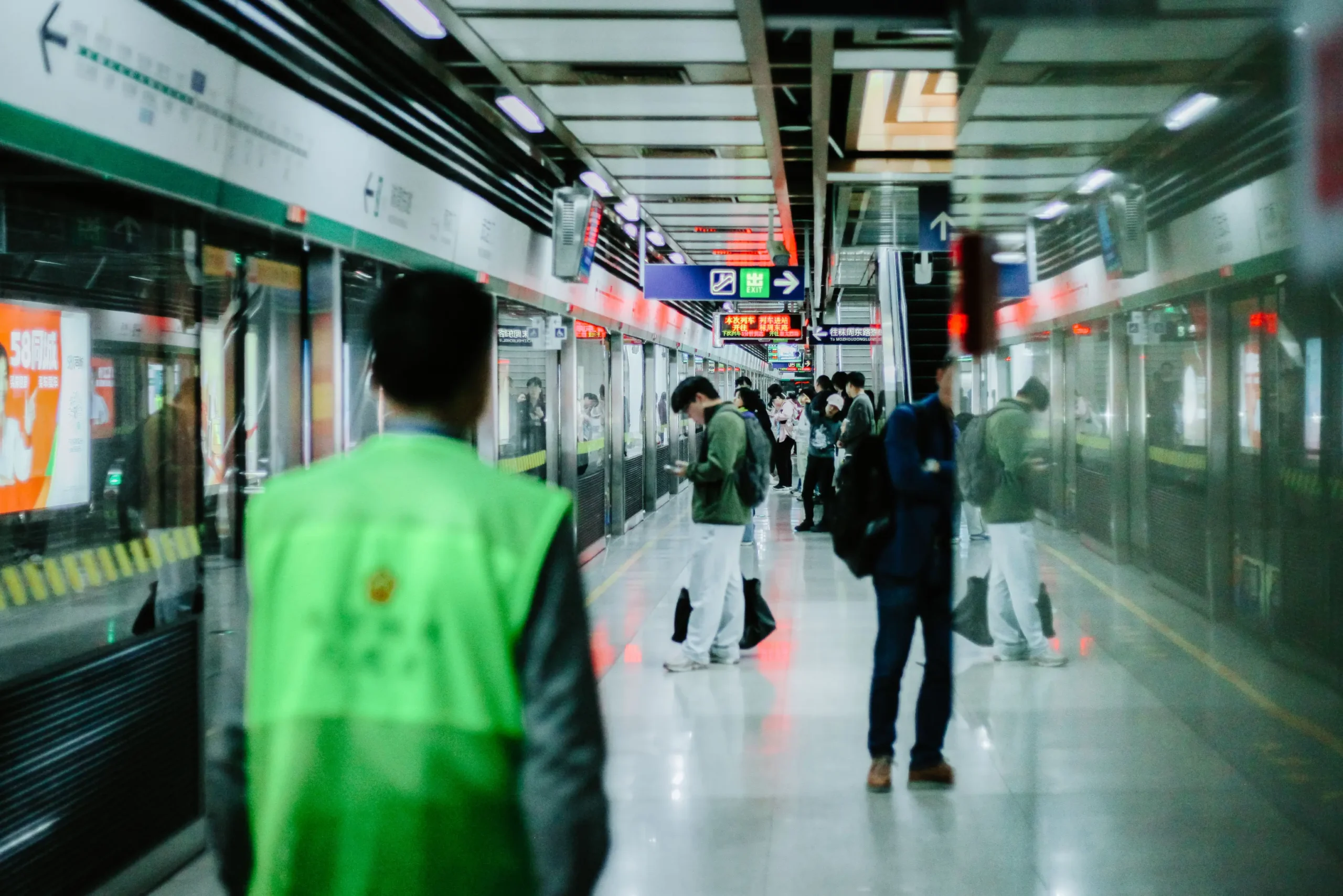 A busy subway platform shows commuters waiting for a train behind glass safety doors, with a station employee in a bright green vest blurred in the foreground. Signs and digital displays hang from the ceiling, casting red and white reflections across the polished floor.