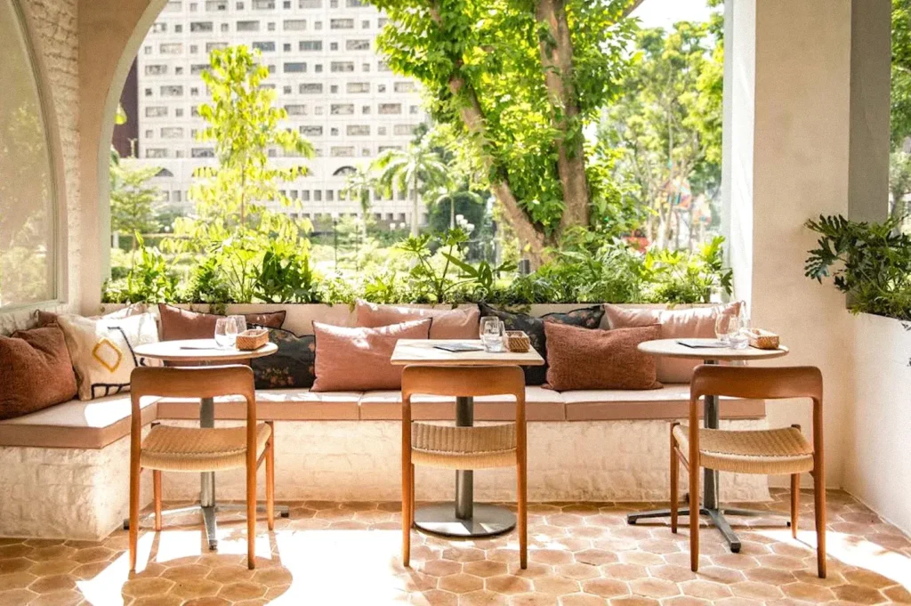 Bright outdoor dining area with round tables, wooden chairs, cushioned bench seating, and lush greenery overlooking a city building.