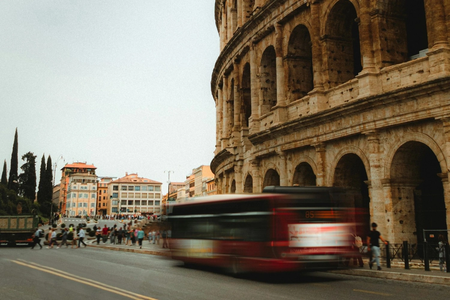 A side view of the ancient, multi-tiered stone arches of the Roman Colosseum towering over a city street on an overcast day. In the foreground, a red city bus is captured in a motion blur as it drives along the asphalt road.