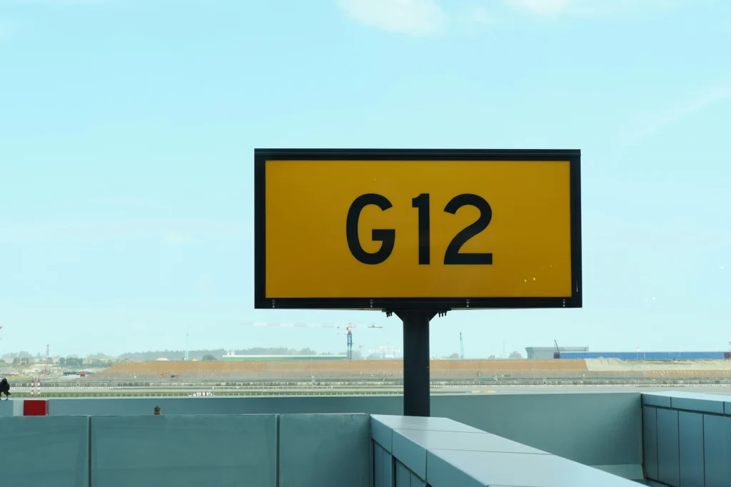 A large, rectangular yellow sign with a black border displays the text "G12" in bold black font. The sign is mounted on a black pole against a pale blue sky, with an airfield and construction cranes visible in the distant background.