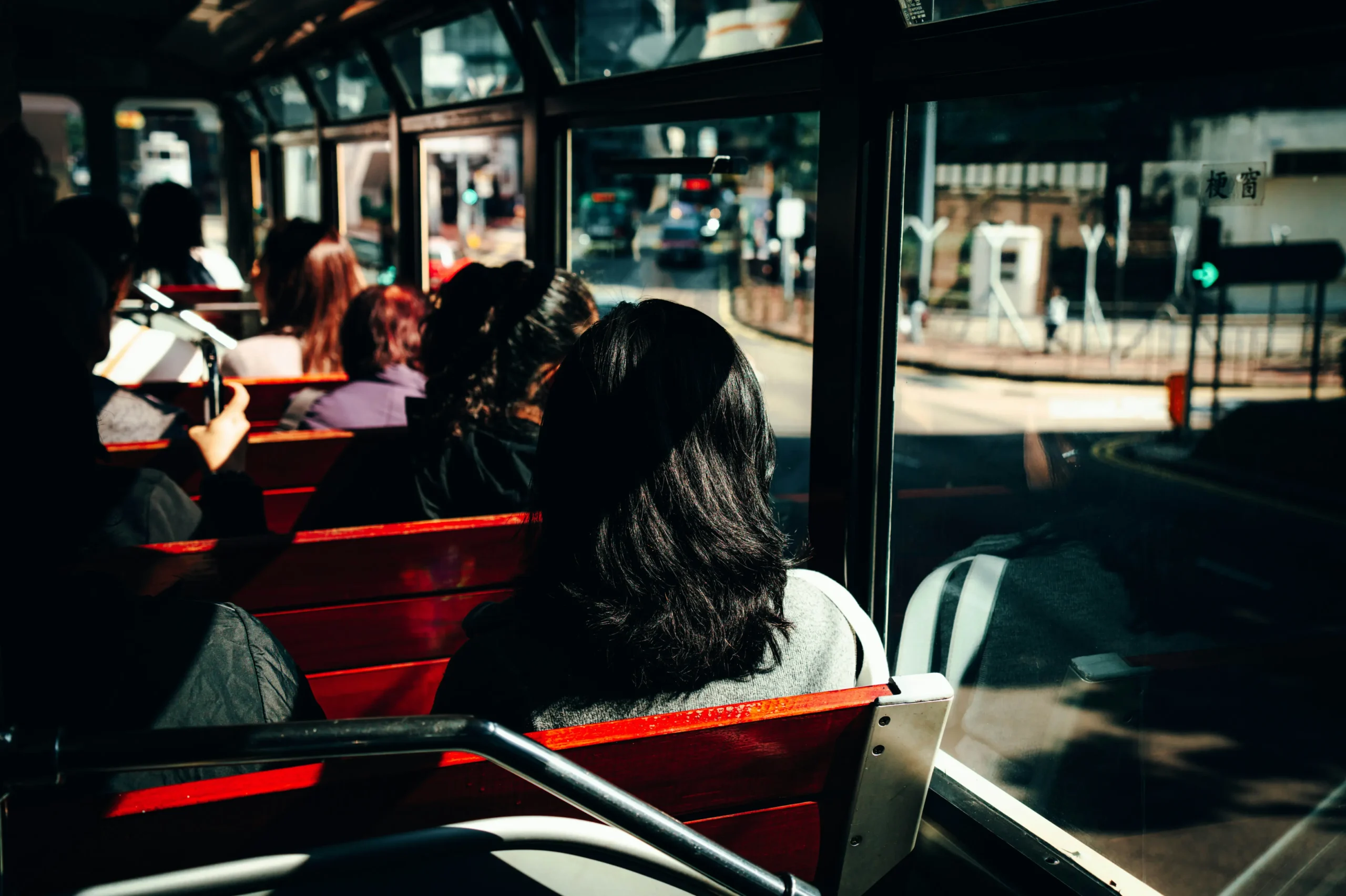 An interior view looking forward from the back of a vintage tram or bus, showing passengers seated on wooden benches with red backs. The sunlight streams through the large glass windows on the right, casting sharp shadows and illuminating a bustling city street outside.