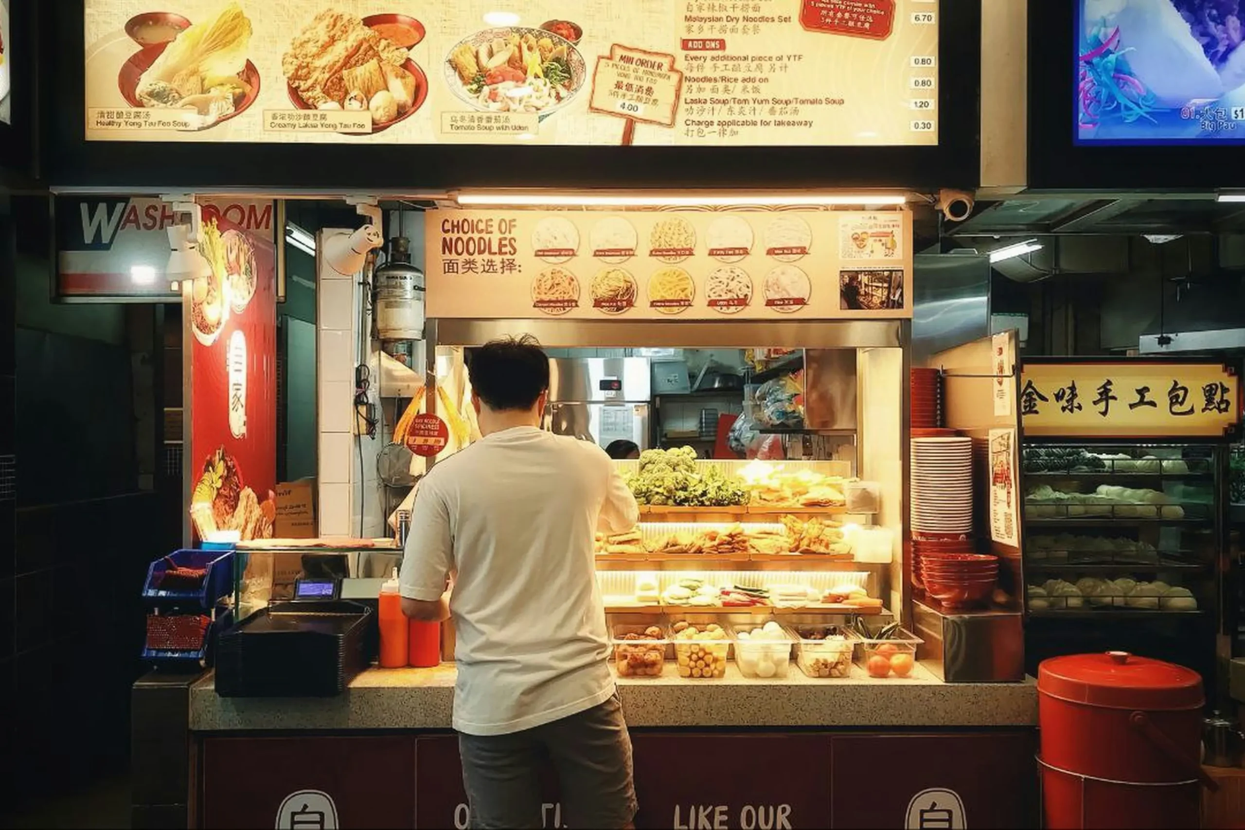 A person in a white shirt and shorts stands facing a brightly lit hawker stall counter that displays an array of fresh ingredients and food items. Above the counter, illuminated menus and signs show food options including "Choice of Noodles" alongside pictures of various dishes.